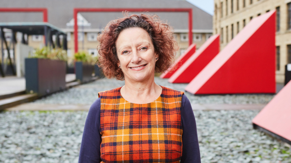 A headshot of Julia Skelton. Julia is a white, middle aged woman with brown eyes and dark red curly chin length hair. She is wearing a yellow and orange tartan dress. The photo is taken outside in front of a large red vent and an old mill building.
