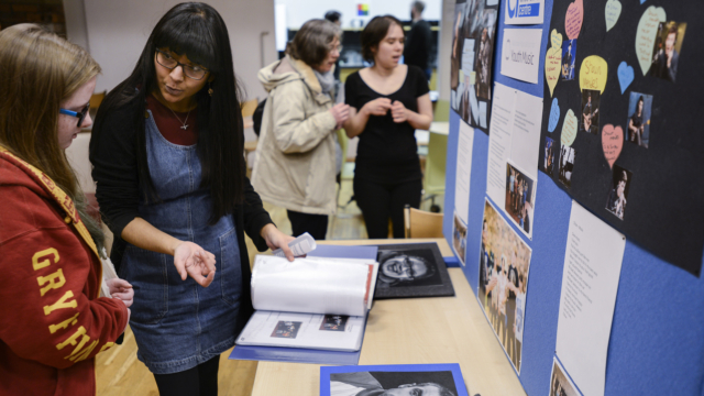 A person with long black hair wearing a denim pinafore shows another person with long hair a portfolio in front of a display at the Academy Open Day