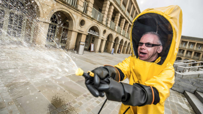 Howard Davies wearing a bright yellow hazmat suit with black latex gloves, pointing and firing a hose