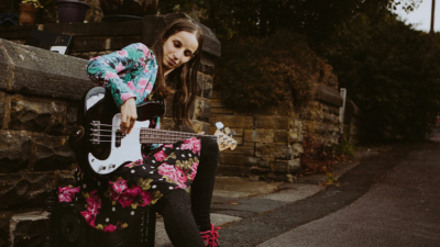 Lorraine Brown sat by a wall wearing a floral skirt and jacket playing an electric bass guitar
