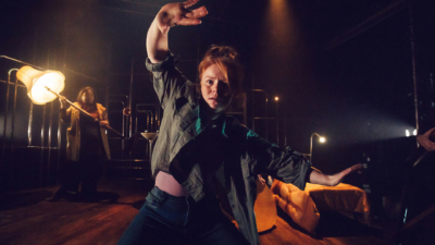 A performer with red hair tied into a bun wearing jeans and a shirt holding their hands up with a shocked expression on their face. A performer in the background is holding a large lamp.