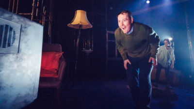 A performer with short hair, a green jumper and dark trousers has a happy, surprised look on their face