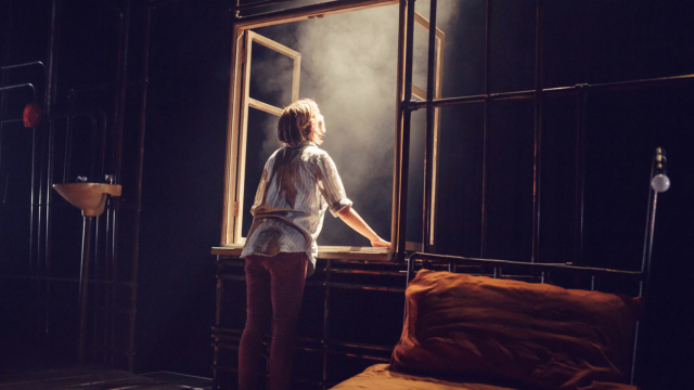 A performer stood beside a bed leaning out of a window frame into bright light and smoke