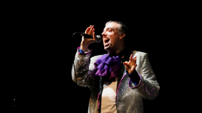 A performer wearing a silver sequin jacket with fancy purple neck ruffle, singing into a microphone