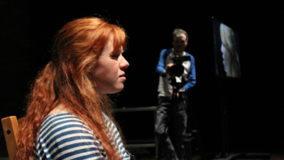 A performer sat in profile with long red hair and a blue and white stripy top, a person in the background is holding a video camera facing the red-headed performer