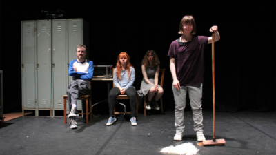 Four performers on stage, three sat in chairs next to a set of lockers and a microwave, one stood holding a broom