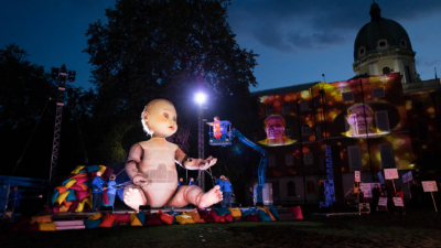 Performer raised in a cherry picker at eye-level with giant model baby that has puppetry arms being moved by a team of puppeteers. In the background are performers holding protest signs and a person's face is projected in lights twice onto a building behind them.