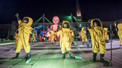 A group of performers wearing hazmat suits, carrying brooms, dancing in front of a giant model of a baby