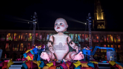 A giant sculpture of a baby with bright blue eyes and with puppetry arms being moved by several puppeteers