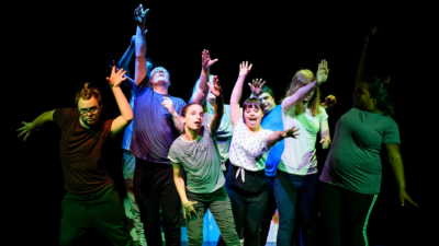 An group of theatre performers with learning disabilities raise their arms on a dark stage