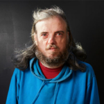 A headshot of Alan Clay. Alan is a white middle-aged man with grey shoulder length hair and a grey beard. He is wearing a red t-shirt with a blue hoodie over the top. He is pictured against a dark background.
