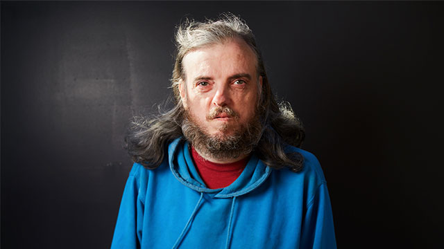 A headshot of Alan Clay. Alan is a white middle-aged man with grey shoulder length hair and a grey beard. He is wearing a red t-shirt with a blue hoodie over the top. He is pictured against a dark background.