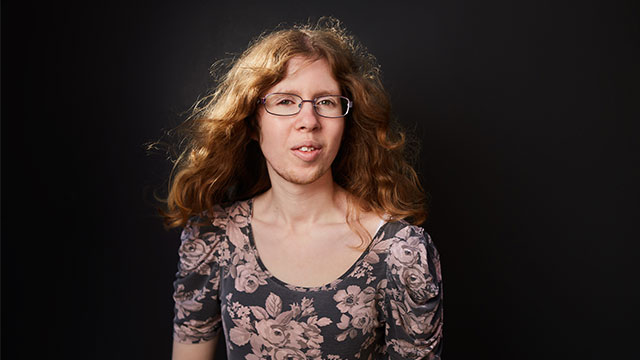 A headshot of Alison Colborne. She is a white woman with shoulder length wavy mousey brown hair. She is wearing dark rimmed rectangle glasses and a flowery top. She is pictured against a dark background.