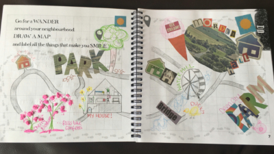 An open notebook with text that reads 'Go for a wander around your neighbourhood. Draw a map and label all the things that make you smile.' with a hand-drawn map, drawings, and a collage of photos and letters stuck onto the pages