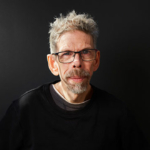 A headshot of Jez Colborne. Jez is a white man with grey short hair and a grey beard. He is wearing dark rimmed, rectangle glasses and a black long sleeve top. He is pictured against a dark background.
