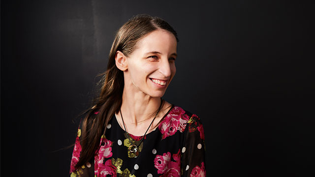A headshot of Lorraine Brown. Lorraine is a white woman with long straight brown hair. She is smiling and looking off towards the side slightly. She is wearing a pink and black floral top. She is pictured against a dark background.