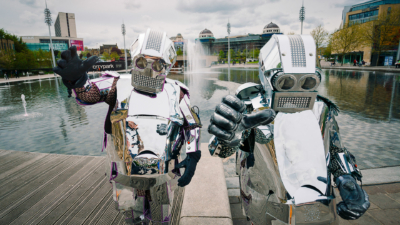 Two people wearing suits made of sheets of silver, resembling robots stood in front of a fountain