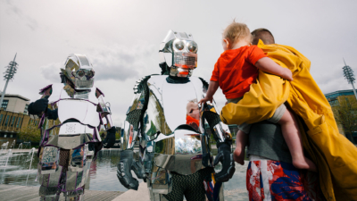 Two people wearing suits made of sheets of silver, resembling robots being approached by a person holding a small child who is reaching out to touch one of the silver people