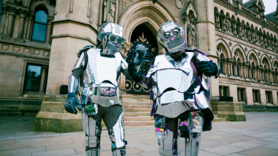 Two people wearing suits made of sheets of silver, resembling robots