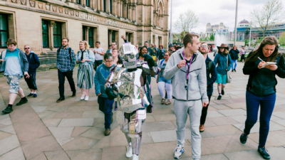 A person wearing a suit made of sheets of silver, resembling a robot, walking with a group of people