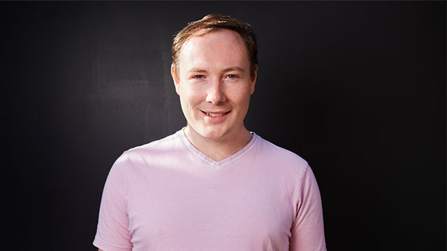 A headshot of Paul Bates. Paul is a white man with short dark blonde hair. He is smiling and wearing a pale pink t-shirt. He is pictured against a dark background.