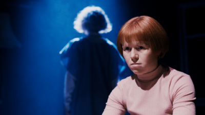 A woman with red hair looks upset in the foreground of the image. In the background is a ghostly figure with her back to the camera lit in blue light.