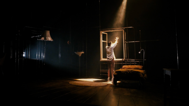A woman in a white shirt and brown trousers has her back to the camera. she is opening a window. A shaft of light is shining through the window.