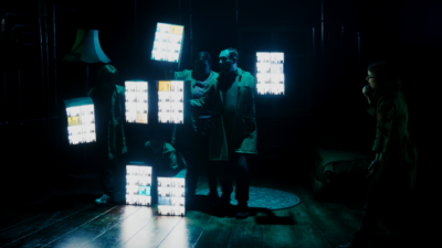 Four people hold seven light boxes to make the shape of a person. A woman to the right of the image appears to be conducting them.