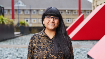A headshot of Bee Skivington. She is an Indian woman in her thirties with dark eyes and long black hair with a fringe. She is wearing dark rimmed glasses and a black and gold shirt dress. The photo is taken outside in front of a large red vent.