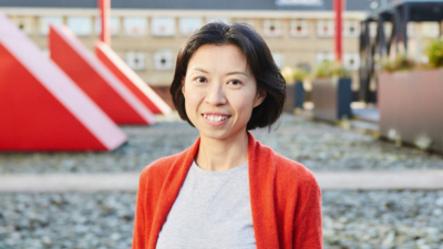 A headshot of Joyce Lee. She is a Chinese woman in her thirties with dark eyes and bobbed black hair. She is wearing a grey t-shirt and a red cardigan. The photo is taken outside in front of a large red vent.