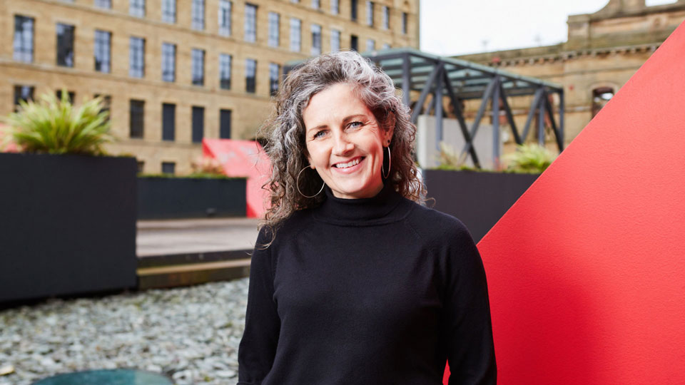A headshot of Maria Thelwell. She is a white middle aged woman with blue eyes and long brown and grey curly hair. She is wearing a black turtle neck jumper and large hoop earrings. The photo is taken outside in front of a large red vent and an old mill building.