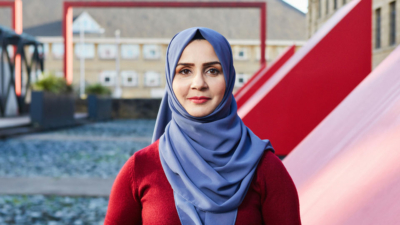 A headshot of Naelah Shahzad. She is a Pakistani woman in her thirties with dark eyes and red lipstick. She is wearing a blue hijab over her hair and a red jumper. The photo is taken outside in front of a large red vent.