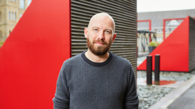 A headshot of Rob Abbey. He is a white man in his thirties with blue eyes, a bald head and a brown beard. He is wearing a smart dark jumper. The photo is taken outside in front of a large red vent.