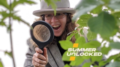 A white woman in a grey blazer and grey bowler hat looks through amagnifying glass pointed just below the camera. She is surrounded by green leaves. The image says the words Summer Unlocked on it.