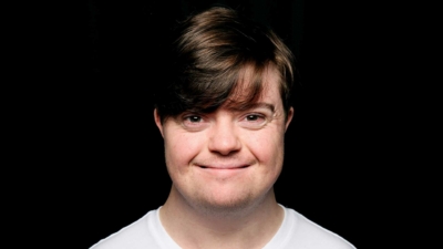 An image of Liam Bairstow - an early thirties white man with downs syndrome. He has dark hair swept across his forehead and is wearing a white t-shirt