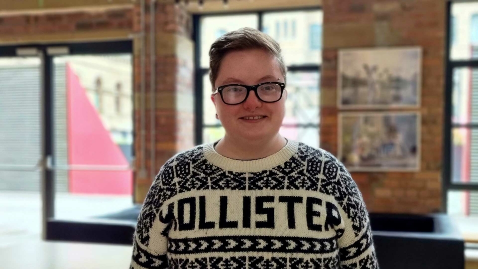 A white mid twenties man stands in a rooms with windows in the background. He has blonde hair, is wearing thick black rimmed glasses and is wearing a knitted Hollister jumper.