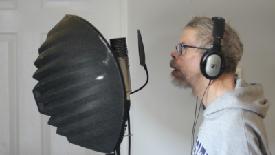 A white man with grey hair and a beard sings into a microphone. He has headphones on and there is a large black dome behind the microphone.