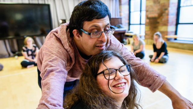 A Pakistani man with short dark hair, glasses and a pink jumper has his arms outstretched around the head of a white woman wearing glasses who has long curly brown hair.