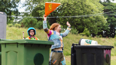 A woman with ginger bobbed hair holds a giant sandwich box in the air. She is about to place it into a black wheelie bin, which is next to a green wheelie bin.