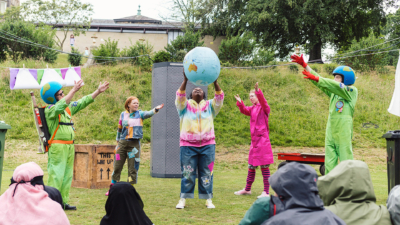 A black woman in a colourful tie dyed hoodie is in the centre of the image holding an inflatable earth globe above her head. Two women and two men stand at either side of her gesturing towards the globe.