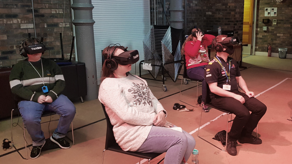 Four people sit on chairs. They each have a virtual reality headset and headphones on.