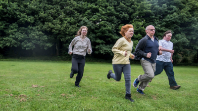 Four people (two women and two men) running through a field with trees behind them.