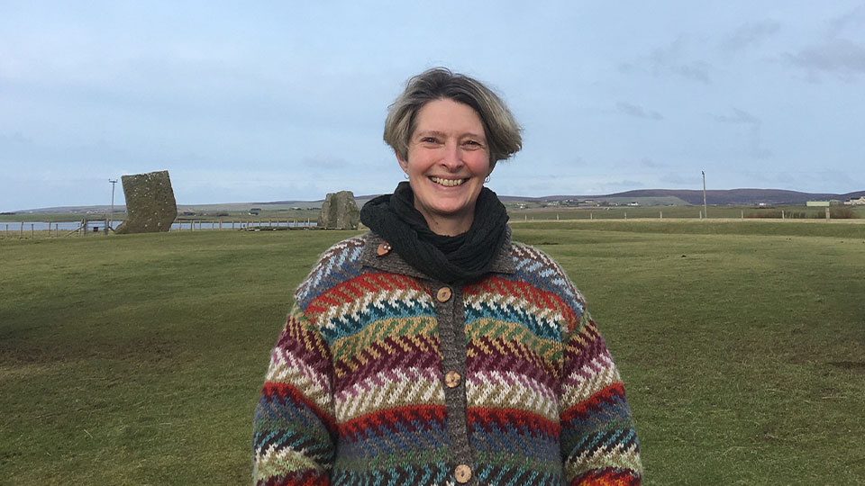 A lady with cheek length mousy hair is stood in a large field with large stone structures in the background. She is smiling and wearing a zig-zag patterned fleece jacket.