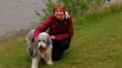 A lady crouched down next to a large scruffy grey and white dog. They are next to a river. The lady has short red hair and is wearing a red and black walking coat.