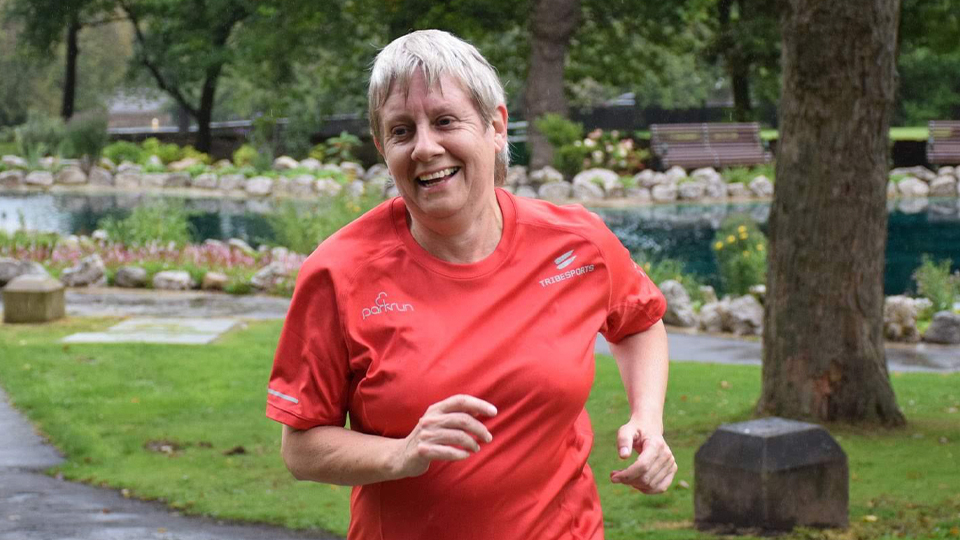 A lady with short grey hair running through a park with a big smile on her face. She is wearing a red running t-shirt.