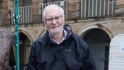 A man with white hair and a short beard. He is wearing a black walking jacket and glasses and is stood in front of a historical building.