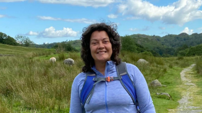 A lady with shoulder length dark hair stood in the countryside with a path leading off behind her to the right of the image. She has a big smile on her face and is wearing a purple walking top and backpack.