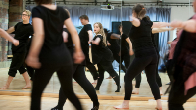 An image of a group of people, all wearing black, and dancing in a studio. The people have their backs to the camera but several people can be seen from the front in a mirror in front of them.