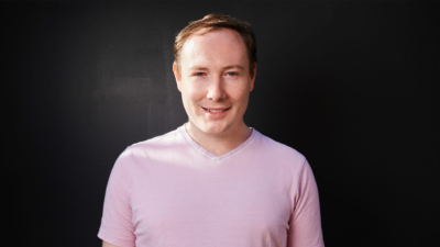 A headshot of Paul Bates. Paul is a white man with short dark blonde hair. He is smiling and wearing a pale pink t-shirt. He is pictured against a dark background.