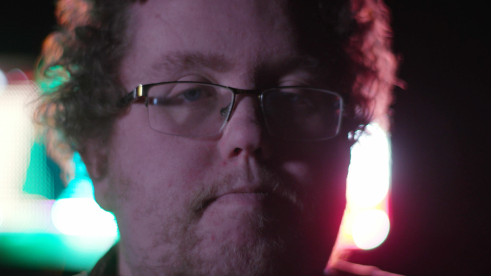 A close up of a mans face. He is white, in his mid thirties and has curly hair, fine rimmed glasses and short stubble. He is back lit by a red and green light.